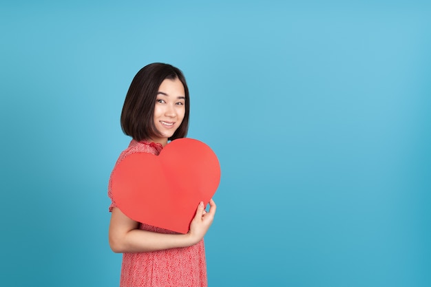 Heureux, souriant jeune femme asiatique en robe rouge tenant grand coeur de papier rouge comme fan