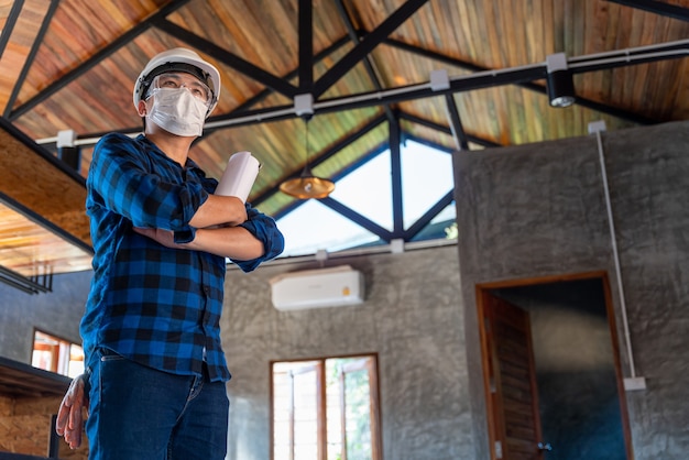 Heureux technicien ingénieur en construction asiatique debout avec les bras croisés en toute confiance après avoir inspecté la structure en bois sous le toit sur le chantier de construction ou sur le chantier d'une maison.