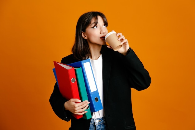 Heureux tenant des dossiers avec une tasse de café jeune belle femme portant une veste noire isolée sur fond orange