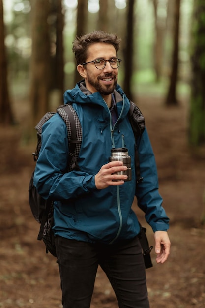 Heureux touriste mâle caucasien du millénaire dans des verres et une veste boire une tasse de café chaud se reposant dans la forêt froide