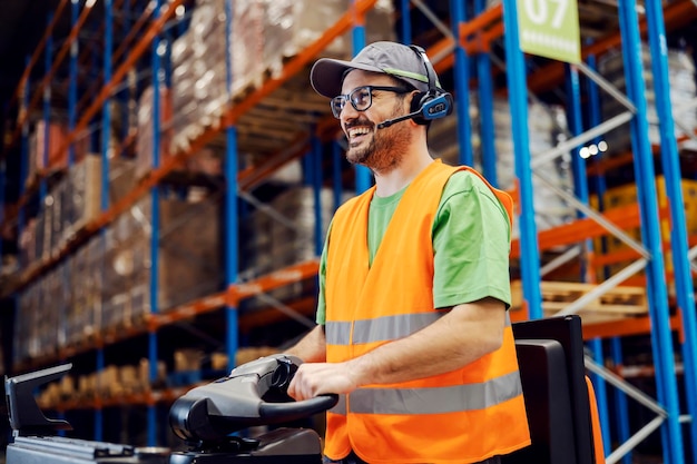 Heureux travailleur du centre de distribution avec casque de sélection vocale conduisant un chariot élévateur