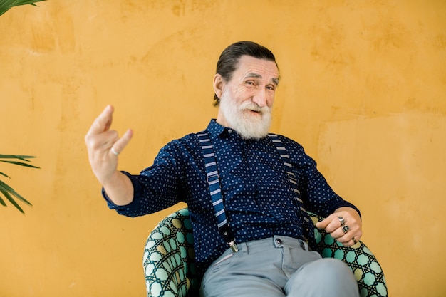 Heureux vieil homme à la barbe grise, portant des vêtements élégants à la mode, posant en studio, assis devant un mur jaune et montrant le rock and roll