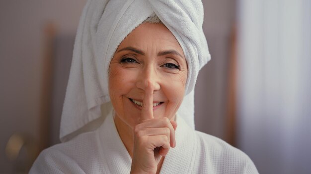 Photo heureux vieilles femmes avec une serviette sur la tête en peignoir regardant la caméra montrant le geste du secret du silence