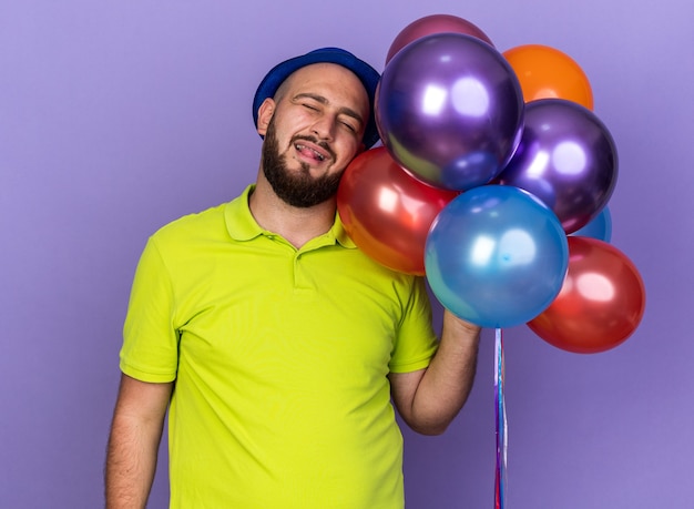 Heureux avec les yeux fermés, jeune homme portant un chapeau de fête tenant des ballons montrant la langue isolée sur le mur bleu