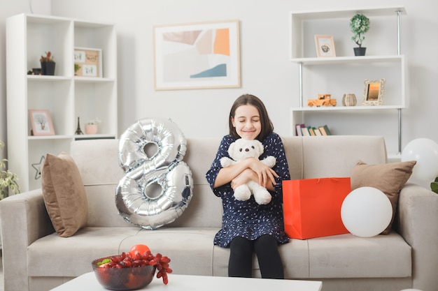 Heureux avec les yeux fermés petite fille le jour de la femme heureuse tenant un ours en peluche assis sur un canapé dans le salon