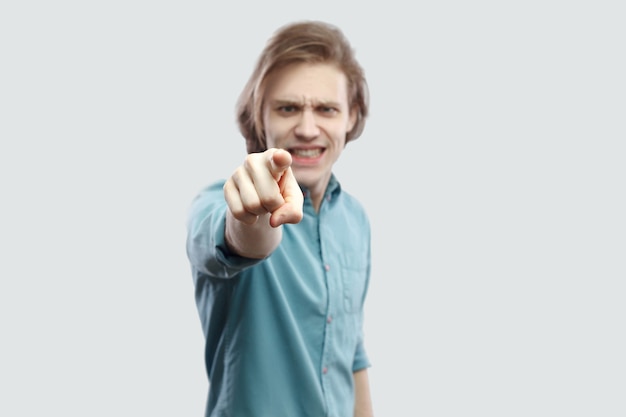 Hey vous. Portrait de blâme sérieux beau jeune homme blond aux cheveux longs en chemise décontractée bleue debout, pointant, alarmant et regardant la caméra. tourné en studio intérieur, isolé sur fond gris clair.