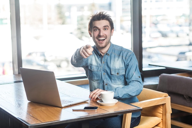 Hey vous! Portrait d'un jeune pigiste barbu heureux et positif en chemise de jean bleu assis dans un café et travaillant sur un ordinateur portable avec un sourire à pleines dents et pointant le doigt vers vous, regardant la caméra. Intérieur