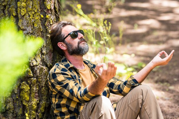 Un hipster adulte barbu médite et se détend en plein air dans les bois de la forêt assis contre un tronc d'arbre et profitant de la nature et de l'amour autour Les hommes profitent de la vie