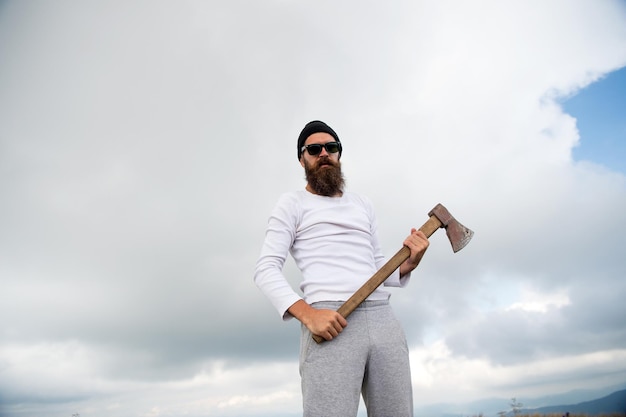 Hipster avec barbe sur un visage strict tient une hache, fond de ciel nuageux. L'homme à la barbe tient une hache tout en se tenant au sommet de la montagne. Hipster a l'air brutal et élégant avec des lunettes de soleil. Conquérant du concept supérieur.