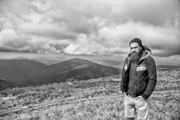 Hipster barbu longue barbe caucasien brutal avec moustache mec mal rasé en veste avec des cheveux élégants se coupe la barbe au sommet d'une montagne venteuse sur un espace de copie de ciel nuageux naturel