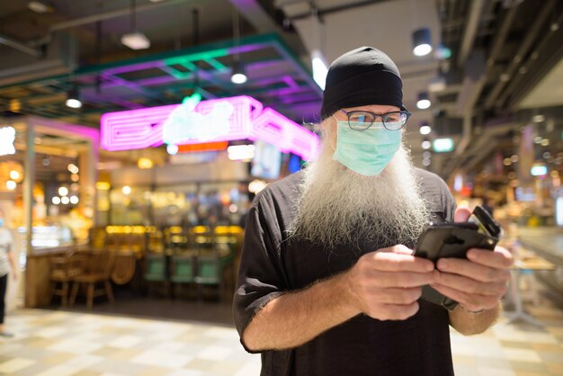 Hipster barbu mature homme utilisant un téléphone avec un masque pour la protection contre l'épidémie de virus corona au centre commercial