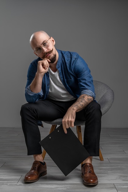 Hipster barbu avec moustache portant des lunettes qui assis sur une chaise en studio dos gris isolé