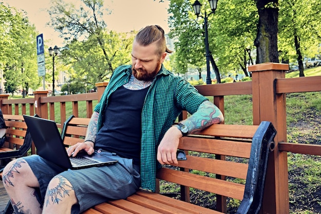 Hipster barbu rousse mâle dans une chemise polaire verte à l'aide d'un ordinateur portable dans un parc.