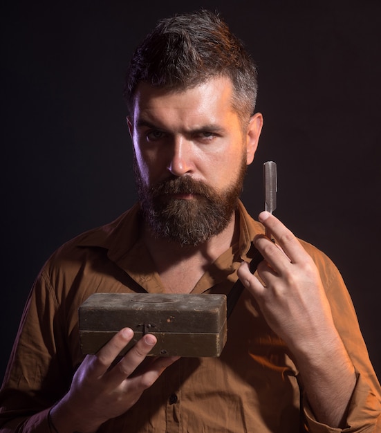 Hipster caucasien brutal homme barbu sérieux avec une longue barbe tient dans les mains un rasoir dangereux en bois