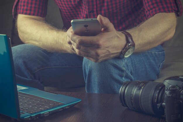 Hipster dans une chemise rouge et un jean bleu est assis avec un smartphone sur le canapé. Notebook et SL