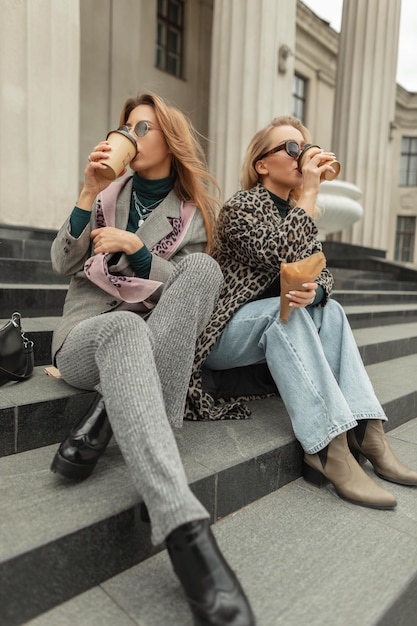 Hipster de deux filles urbaines heureuses de mode dans des vêtements d'automne à la mode élégants avec des chaussures voyage et s'assied sur les marches de la ville boit du café dans des tasses