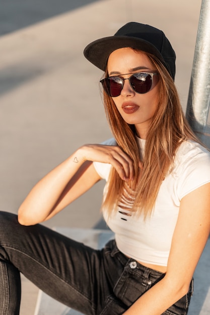 Hipster femme élégante moderne en casquette noire à la mode en T-shirt sexy déchiré en jeans en lunettes de soleil mode se détend près du pilier métallique dans le parking de la ville par une journée ensoleillée. Fille cool urbaine en ville au coucher du soleil.