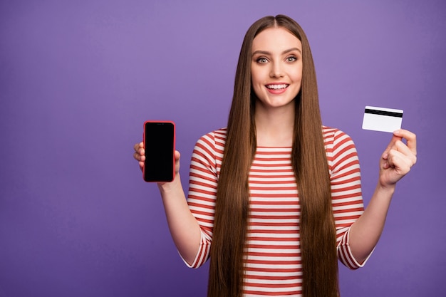 Hipster fille positive tenir la carte de crédit smartphone présente la promotion de la technologie moderne elle paie le service de paiement bancaire facile porter pull pull blanc rayé mur de couleur violet isolé