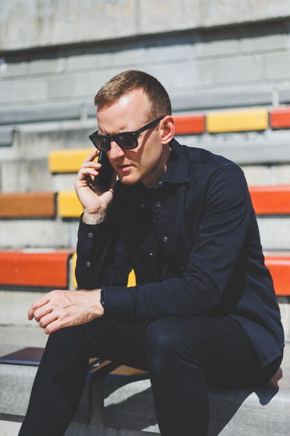 Hipster gai en tenue à la mode souriant tout en se reposant dans un cadre urbain en ville à l'aide d'un téléphone portable sur les loisirs heureux blogueur masculin du Moyen-Orient en lunettes de soleil appréciant le mode de vie de voyage