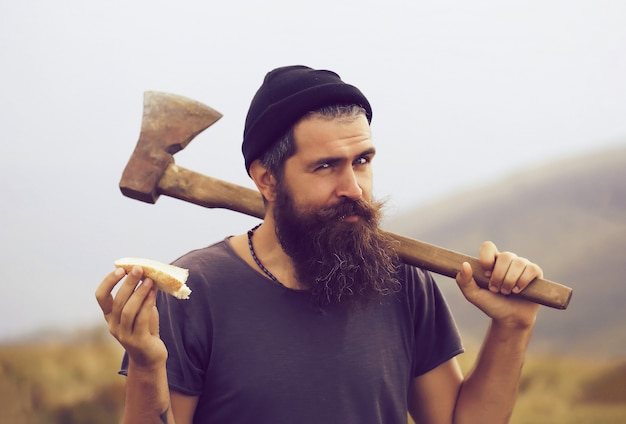 Hipster homme barbu bûcheron avec barbe et moustache avec hache manger du pain au sommet de la montagne