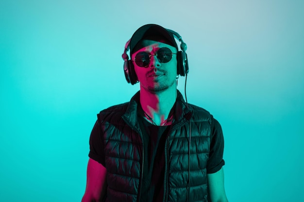 Hipster homme branché élégant en lunettes de soleil à casquette noire et écouteurs en vêtements de mode noirs en studio avec une couleur de lumière bleue et rose