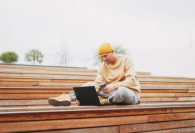 Hipster homme élégant en pigiste de chapeau jaune travaillant sur ordinateur portable dans le parc de la ville de rue