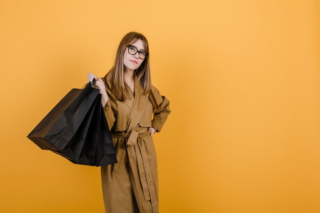 Hipster jeune femme à lunettes et automne trench-coat avec des sacs shopping noirs isolés sur jaune