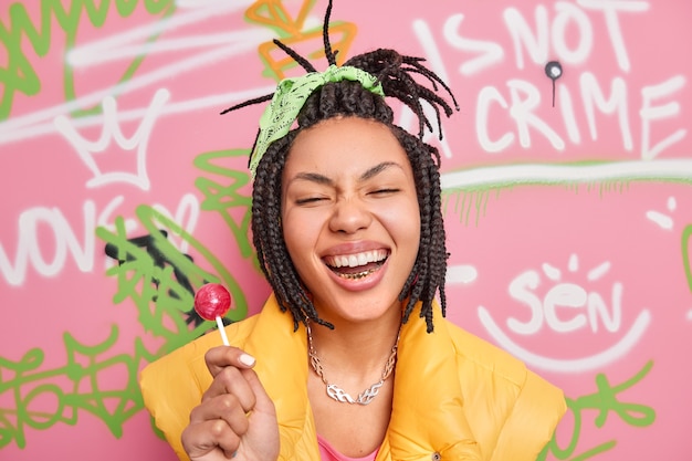 Hipster joyeuse branchée sourit largement tient la sucette s'amuse avec des adolescents du même âge porte un gilet jaune pose contre un mur de graffitis colorés