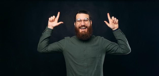 Hipster mâle barbu extatique pointe vers le haut avec les deux mains tout en souriant Panorama tourné en studio sur fond noir