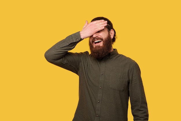 Hipster mâle barbu tient sa paume sur le visage tout en souriant Studio tourné sur fond jaune