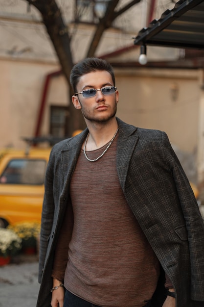 Hipster à la mode jeune bel homme modèle avec des vêtements élégants élégants avec des lunettes de soleil manteau à la mode et sweat-shirt promenades dans la ville