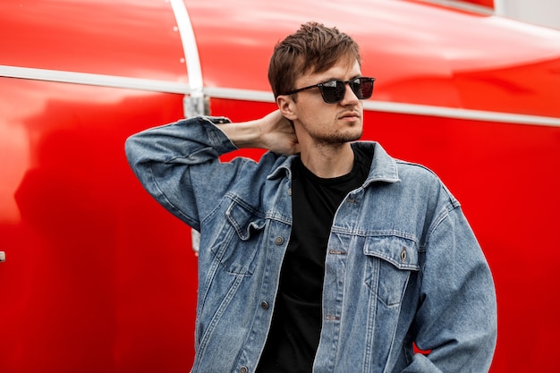 Hipster à la mode jeune homme posant près du mur rouge en métal moderne. Modèle de beau mec dans des vêtements en denim à la mode dans des lunettes de soleil élégantes bénéficie de vacances dans la ville un jour de printemps. Style de rue américain.