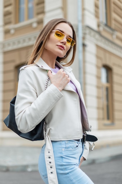 Hipster modèle belle femme à la mode avec des lunettes de soleil jaunes de mode en cuir blanc en cuir avec un jean et un sac se promène dans la rue