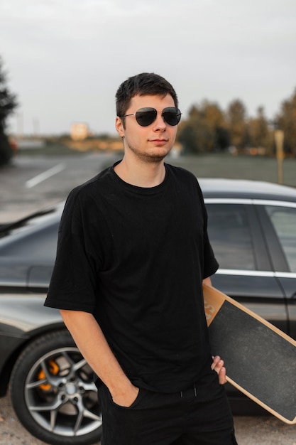 Hipster modèle homme cool en t-shirt noir fashion avec longboard se tient près d'une voiture à l'extérieur