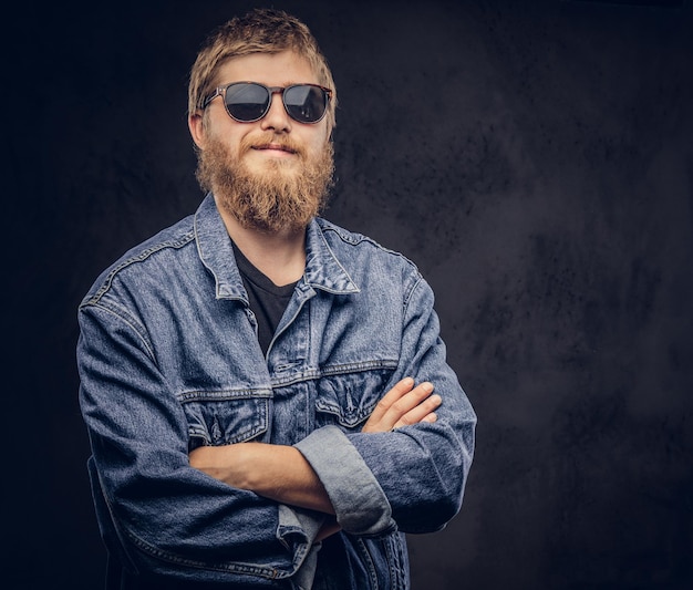 Un hipster pensif portant des lunettes de soleil vêtu d'une veste en jean posant avec les bras croisés. Isolé sur un fond sombre.