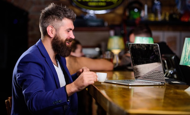 Hipster pigiste travail carnet de blog en ligne Surfer sur Internet Emploi en ligne Homme d'affaires barbu assis pub avec ordinateur portable et tasse de café Avantages indépendants Le gestionnaire travaille en ligne tout en savourant un café