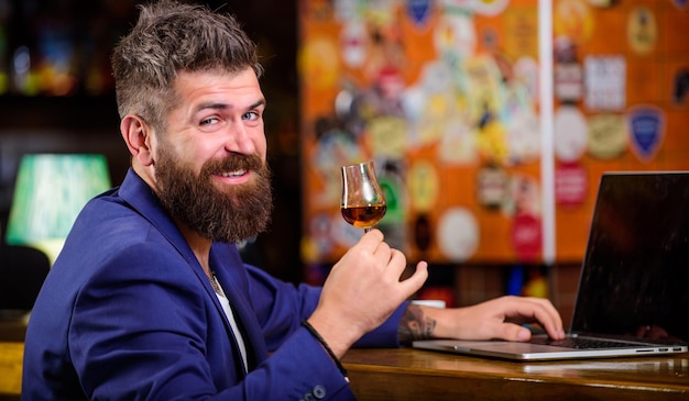 Hipster pigiste travail en ligne boire du cognac Homme barbu homme d'affaires bar comptoir ordinateur portable cognac Agréable se détendre homme d'affaires prospère Célébrer le succès Homme d'affaires surfer sur Internet boire de l'alcool