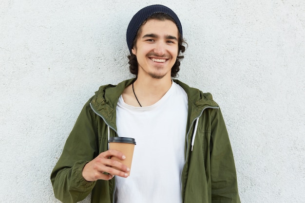 Hipster positif dans un élégant couvre-chef noir, un t-shirt blanc et un anorak, détient une tasse de café en papier, bénéficie de temps libre