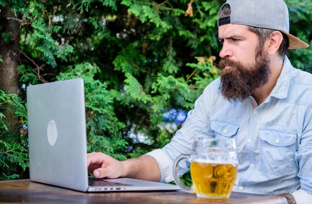Hipster se détendre s'asseoir sur la terrasse avec de la bière Un pigiste hipster barbu profite de la fin de la journée de travail avec une chope de bière La bière l'aide à se détendre après une dure journée Loisirs d'homme brutal avec de la bière et un ordinateur portable Enfin vendredi