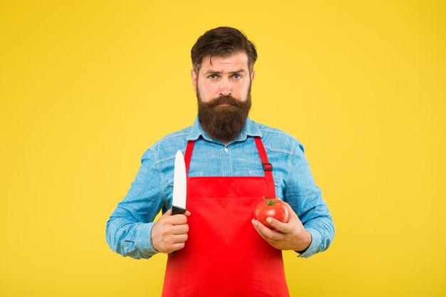 Un hipster sérieux tient une tomate et un couteau pour cuisiner une recette de légumes frais pour une alimentation saine, végétarienne.