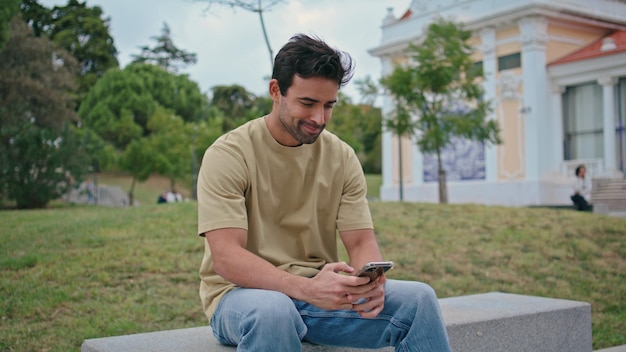 Un hipster souriant envoie des messages sur son téléphone portable sur un banc urbain.