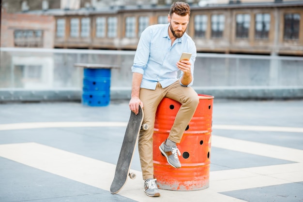 Hipster utilisant un téléphone assis avec une planche à roulettes sur l'aire de jeux sur le toit du bâtiment industriel