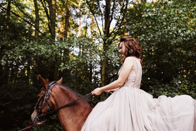 Histoire d'amour et mariage près de la rivière dans un champ au coucher du soleil avec cheval brun. La mariée en robe aérée est de la couleur de la rose poussiéreuse. Robe beige à paillettes.