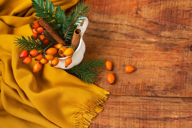 Hiver, ambiance chaleureuse. Une composition avec une tasse avec des branches d'épinette, des églantiers et une écharpe jaune sur un fond en bois