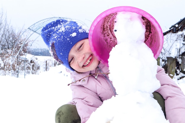 L'hiver est un moment de jeu et de plaisir une fille souriante de huit ans dans une veste pastel et une casquette bleue fait un bonhomme de neige dans la cour en gros plan