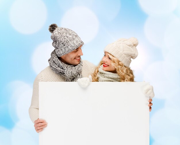 hiver, vacances, noël, publicité et concept de personnes - couple souriant en vêtements d'hiver avec panneau d'affichage blanc vierge sur fond de lumières bleues