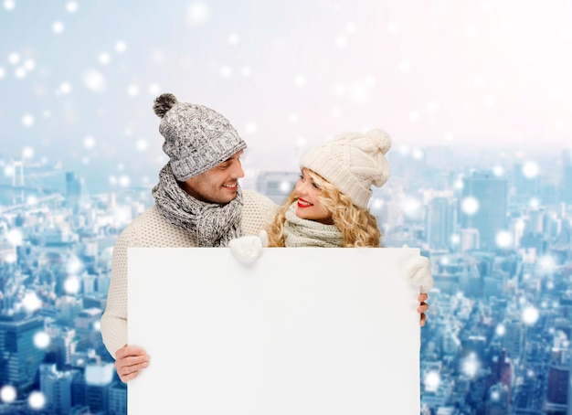 hiver, vacances, noël, publicité et concept de personnes - couple souriant en vêtements d'hiver avec panneau d'affichage blanc vierge sur fond de ville enneigée