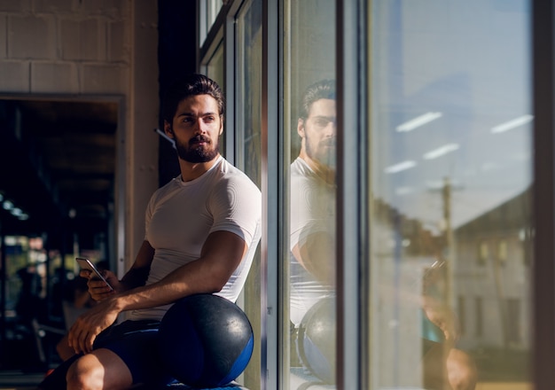 Homme actif sportif assis près de la fenêtre dans la salle de gym avec mobile à la main et grosse balle à côté de lui et à la recherche de loin.