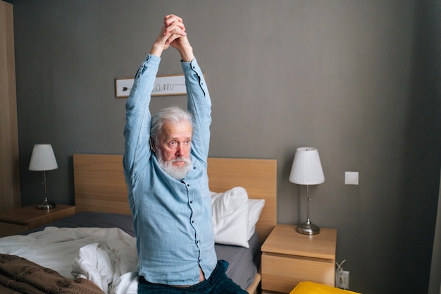 Homme adulte âgé se réchauffant et s'étirant les mains assis sur le lit le matin après s'être réveillé à la maison. Bel homme mûr barbu souffrant de tension musculaire après son réveil.