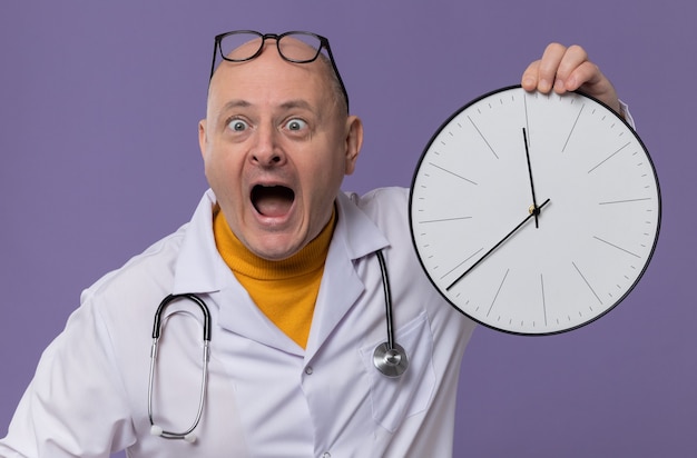 Homme adulte anxieux avec des lunettes en uniforme de médecin avec stéthoscope tenant une horloge regardant de côté